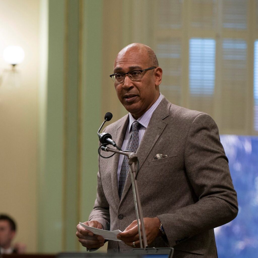 California Assemblymember Chris Holden, pictured in a brown seat speaking into a microphone, has authored a bill that would ban police from lying to minors during interrogations