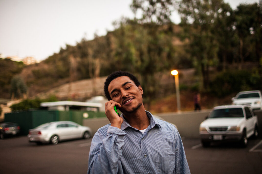Terrick Bakhit, a young Black and Mexican man wearing a blue buttoned shirt, smiles in a parking lot while talking on the phone with his girlfriend.