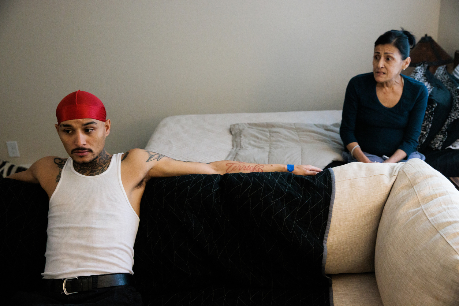 A man wearing a red durag and a white tank top sits on a couch while speaking with his mother, who’s sitting on a bed behind him.