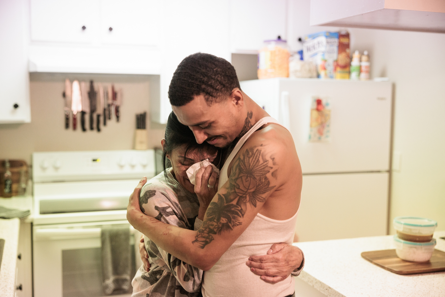 A woman with a tissue to her face cries while being hugged by her son, a man with tattoos and a white tank top.