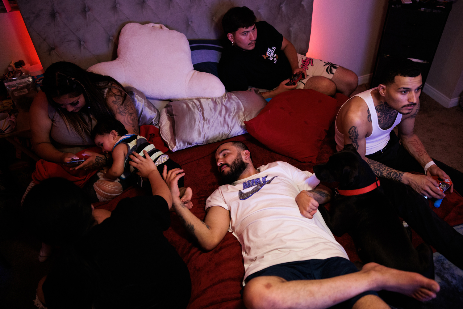 A man wearing a white and blue Nike shirt relaxes on a bed with his relatives. He rests his left hand, which is paralyzed from a past stroke, on his stomach.