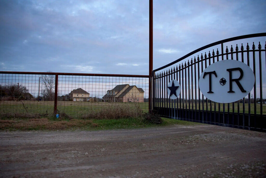 A photo of a gate to the The Thompson’s Residential Treatment Center in Greenville on Wednesday, Feb. 5, 2025. The arched gate has metal bars with the initials "T-R" on it. Several building are seen to the left of the gate behind a metal fence. Photo by Emil T. Lippe for The Texas Tribune.