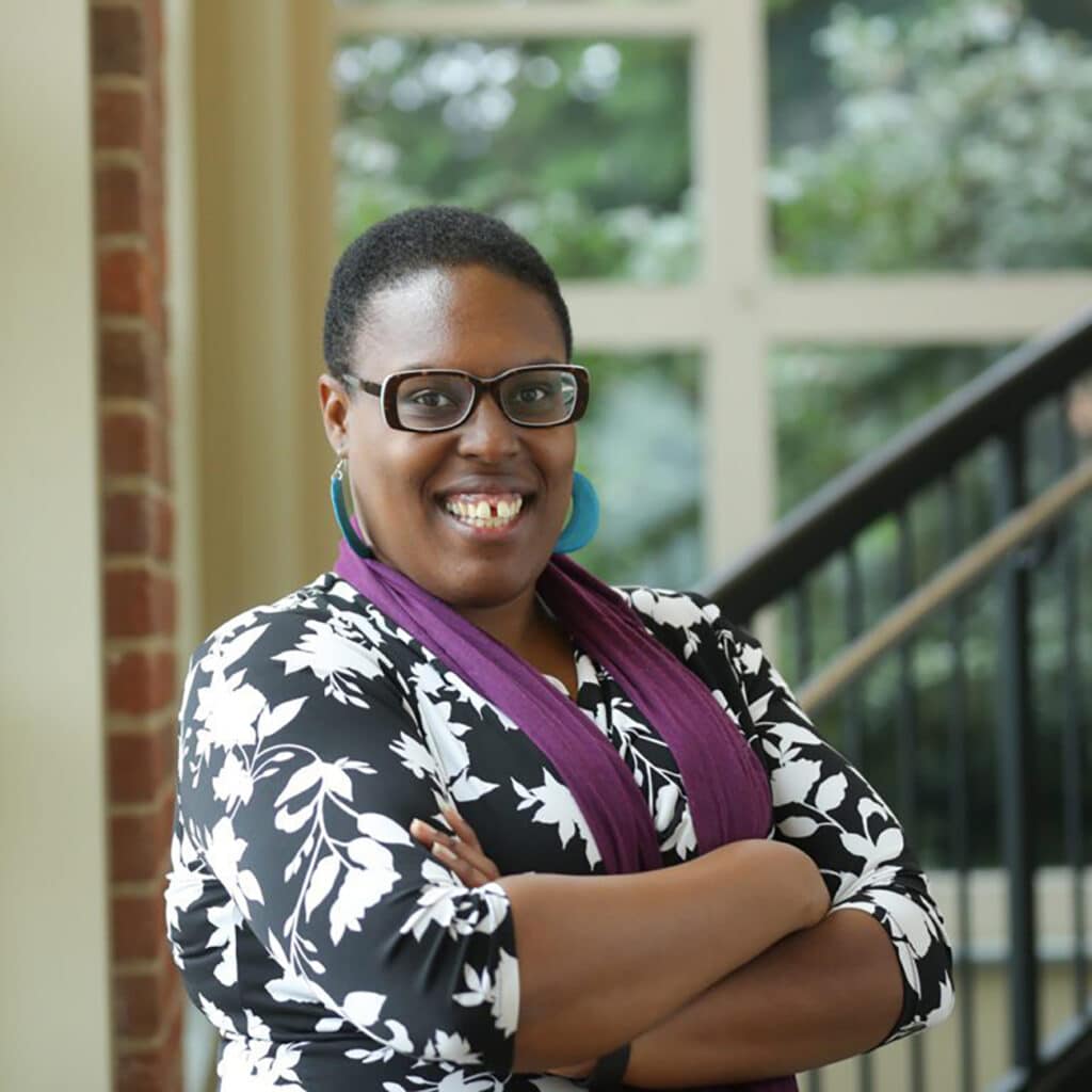 Smiling woman with glasses, her arms crossed over her chest, with a blurred window and stair railing in the background.