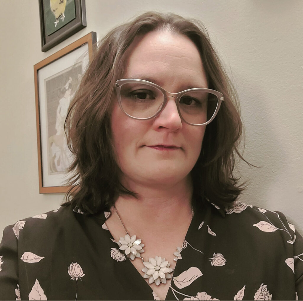 A photo of a woman wearing large glasses with shoulder length hair. Two framed pictures hang on the wall behind her.