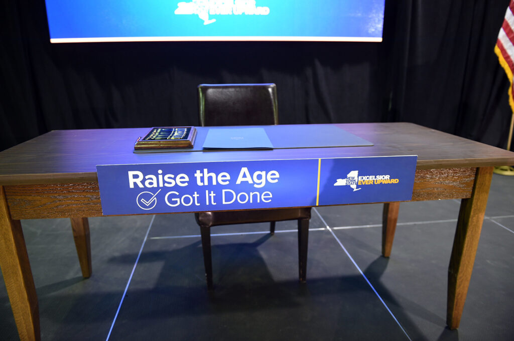 A long, empty desk with microphone and a screen behind it. A sign on the desk reads Raise the Age, got it done.