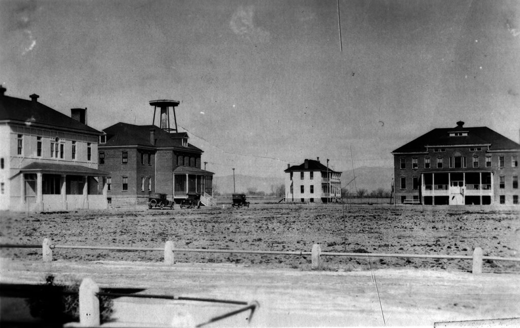 A black and white photo of Teller school in Grand Junction, showing four buildings with Old West style porches. Two old fashioned cars are parked in front of one, possibly Model Ts.