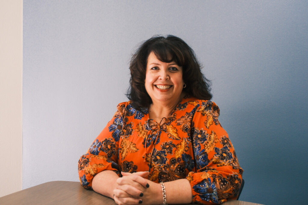Melissa Lloyd has curled brown hair and is sitting at a table with her hands folded. She is smiling, and wearing an orange blouse with blue flowers and brown leaves printed on it.