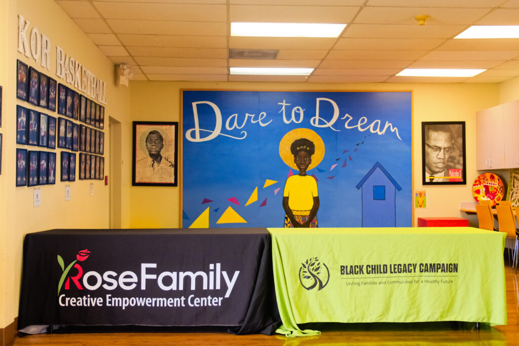 Two conference tables are covered in sheets, one printed with "Rose Family Creative Empowerment Center," and the other one printed with "Black Child Legacy Campaign." Behind it is a blue mural depicting a Black girl in a yellow shirt with the words "Dare to Dream" painted across the top.