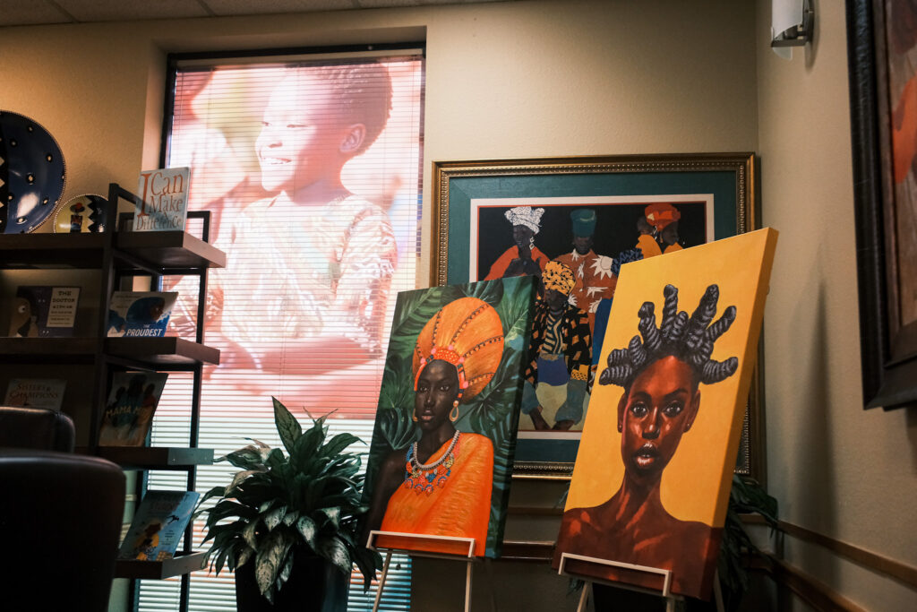 Two paintings depicting Black women rest on stands inside a beige waiting room. Off to the left, a shelf displays children's books.
